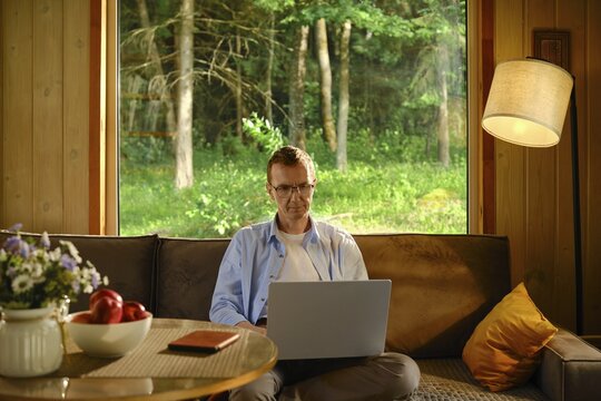Middle-aged man sits on couch in wooden cabin. He focuses on laptop while making online shopping and ordering delivery of goods. Red apples and notebook rest on table nearby