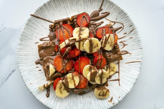 Close up top down view massive, high-calorie waffle topped with strawberries, bananas, and chocolate at an outdoor cafe. A delicious and indulgent big portion sweet treat