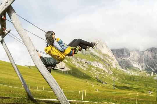 Excited woman with backpack swinging on wooden playground swing lush green hills steep mountain peak misty background Seceda Dolomites Italy summer travel adventure lifestyle