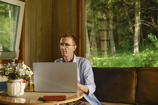 Middle-aged man sits at table using laptop inside wooden cabin and focused on online learning. Sunlight comes through window, casting light on wooden walls. Green trees are visible outside. Small vase with flowers sits on table