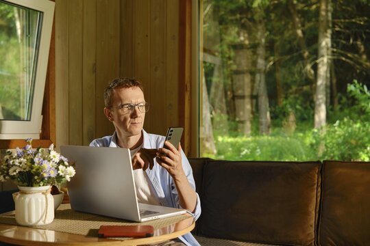 Middle-aged nomad sits at table in wooden cabin using laptop and smartphone. Sunlight shines through windows. Green trees visible outside. Scene shows focus on work amidst natural surroundings