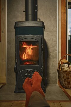 Feet in red socks rest in front of wood stove with flames. Basket nearby shows logs. Room has wooden walls. Light from fire offers simple warmth at night