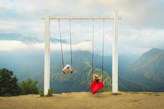 Couple riding giant swing above mountain valley with forest, clouds and panoramic landscape, symbolizing freedom, romance, adventure, wellness and dream travel