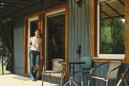 Relaxed middle-aged woman stands on the porch of wooden cabin holding drink. There are two chairs and a small table visible on the wooden deck. It is afternoon