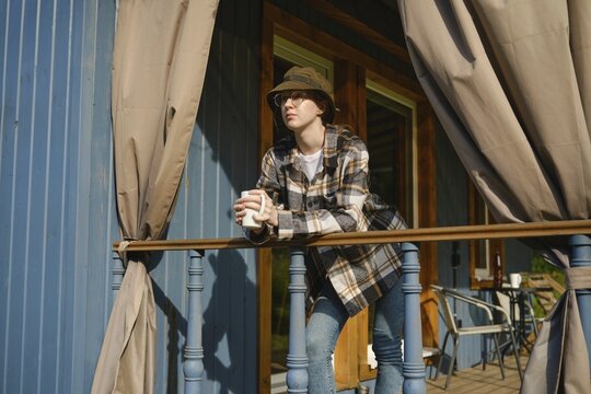Young woman stands on terrace of wooden cabin leaning on railing holding cup of tea. She wears checkered shirt and hat