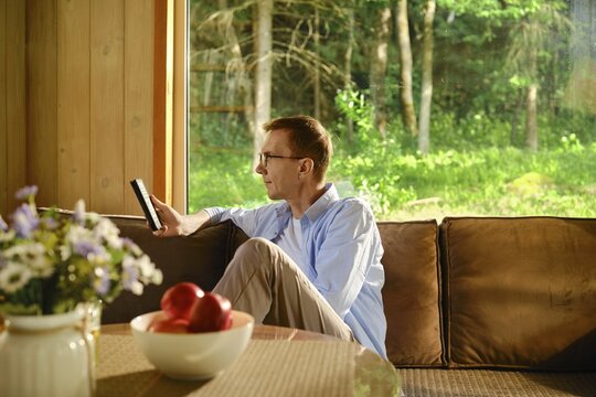Retired man sits on couch in living room in wooden cabin holding electronic boor in hand and reading. Calm and leisurely retirement life away from noisy city and bustle