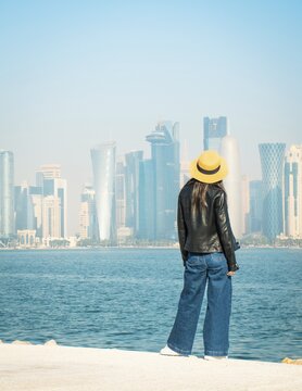 Woman wear casual clothing yellow hat overlooking Doha West Bay skyline. Visible landmarks: Burj Doha, Tornado Tower, Palm Tower, Al Bidda Tower, Qatar Petroleum district. Blue sea background