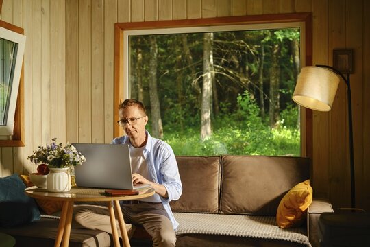 Middle-aged man sits at small round table with laptop in living room in wooden cabin. Sunlight enters through large window. Green trees are seen outside