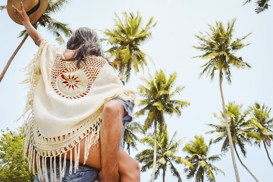 Happy young couple piggyback walking having fun on sandy pathway under tall coconut palm trees generic tropical island beach resort sunny summer day