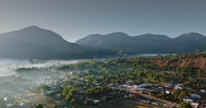 Lombok island in Indonesia, Sembalun village nestled in a lush green valley. Surrounded by towering mountains and shrouded in a thick layer of morning fog illuminated by the warm sunrise light