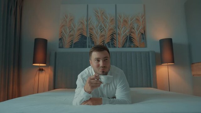 A young man royally drinks tea in a sleeping bed with a contented and dreamy expression on his face.