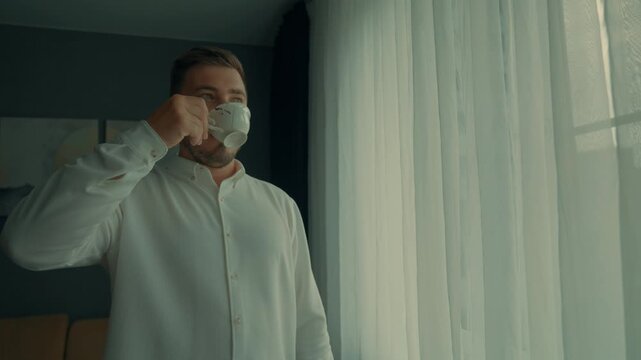 A man in a white shirt drinks coffee or tea before the start of the working day in the morning and looks out the window.