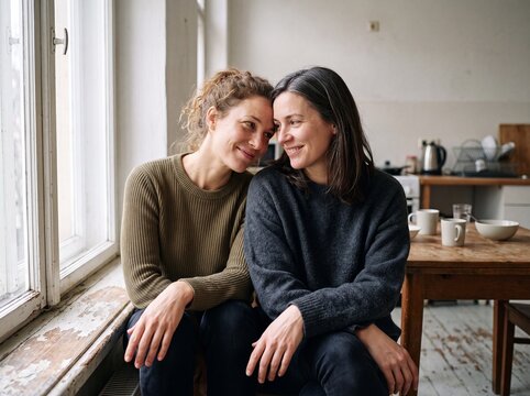 Zwei l&auml;chelnde Frauen sitzen eng beieinander auf der Fensterbank einer gem&uuml;tlichen K&uuml;che und teilen einen vertrauten Moment