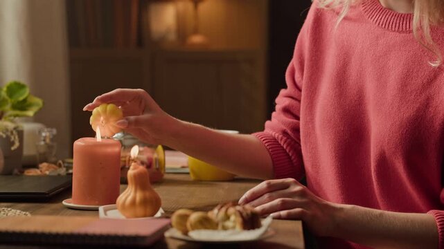 Woman lighting small pumpkin candle from another burning candle wick. Footage is ideal for autumn seasonal marketing, home decor advertisements, and holiday lifestyle content