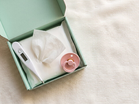 A mint-colored box containing paper tissues, a digital thermometer, and a pink pacifier, resting on a white knitted throw.
