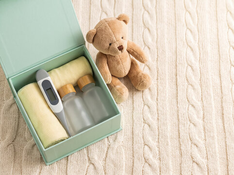 An open box containing bandages, a thermometer, small jars, and a teddy bear, resting on a knitted throw.
