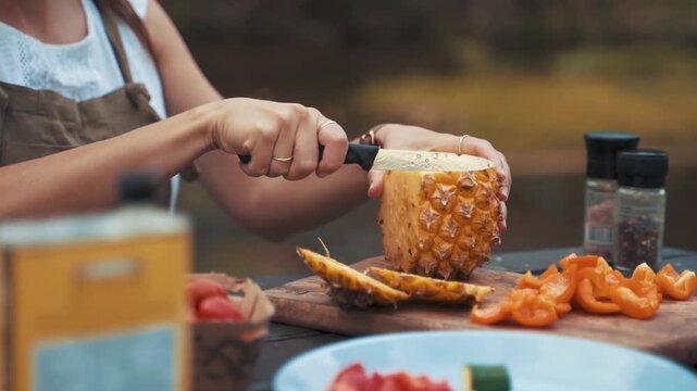 Pineapple stands on a wooden cutting board while a person uses a sharp knife to slice the fruit into wedges as other prepped ingredients and seasoning bottles wait nearby for the final food assembly.