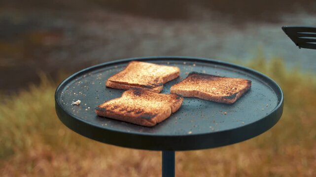 Slices of bread roast slowly on a circular black camping stove positioned outdoors near a grassy field while the morning sun warms the crisp air and creates a serene environment for breakfast time.