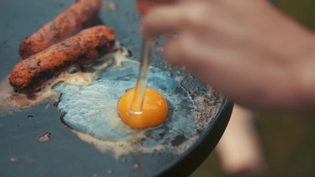 Egg cracked onto a hot cast iron griddle with grilled sausages alongside it is being prepared for a quick breakfast by someone outdoors while enjoying the morning air in a calm and peaceful setting.