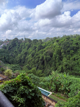 A scenic view of lush green hills under a partly cloudy sky. Tropical vegetation thrives on the hillside, creating a vibrant natural landscape. A swimming pool is visible in the foreground.