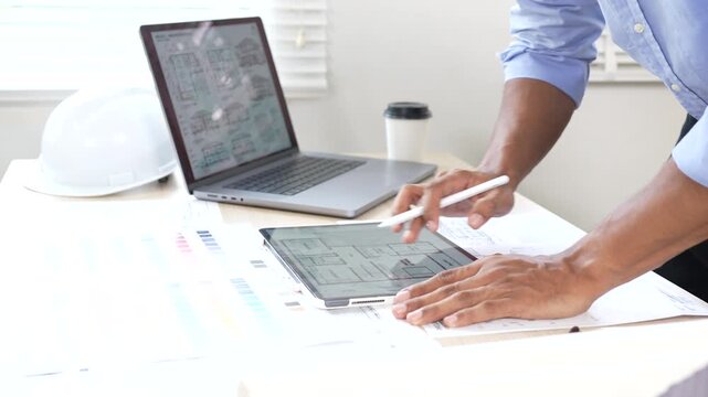 Architect reviewing construction plans at a modern office desk with blueprints, laptop, and engineering tools for a building project.