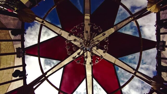 Circular architectural canopy features alternating translucent red and blue roof panels surrounding a suspended floral chandelier beneath an open sky with clouds, static shot.