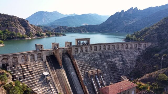 Panoramic view of Oliana dam in Lleida, Catalonia, with stepped concrete spillway, arched gallery crest, powerhouse, penstock pipes and pre-Pyrenean mountains framing a turquoise reservoir