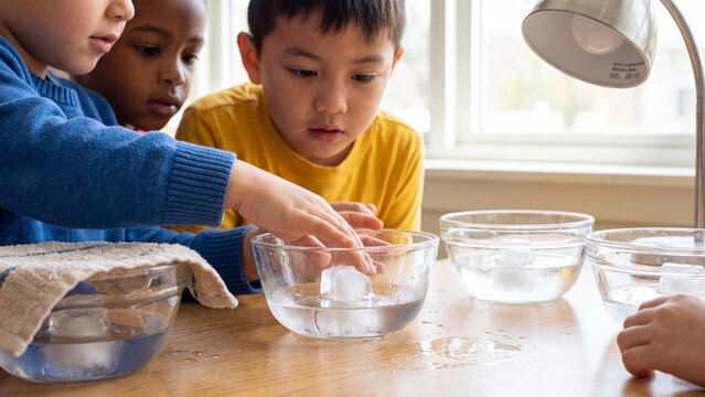 Children observe ice cubes melting in glass bowls while participating in a collaborative science experiment inside a brightly lit classroom setting.