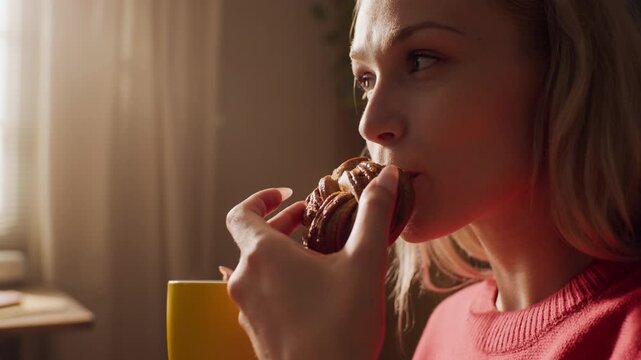 Young adult Caucasian woman holding yellow mug and eating sweet pastry at home. Footage is ideal for breakfast food marketing, bakery promotions, or morning routine lifestyle advertisements