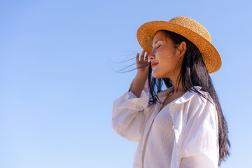Side Profile Portrait of Young Woman Wearing Straw Hat Against Clear Blue Sky, Summer Vacation, Beach Travel, Relaxation, and Lifestyle © kwanchanok