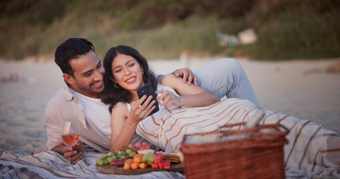 Couple, picnic and relax on beach with phone, wine and fruit platter with online search for bonding. Happy man, woman and scroll app, internet and charcuterie with drink for break by coast for love