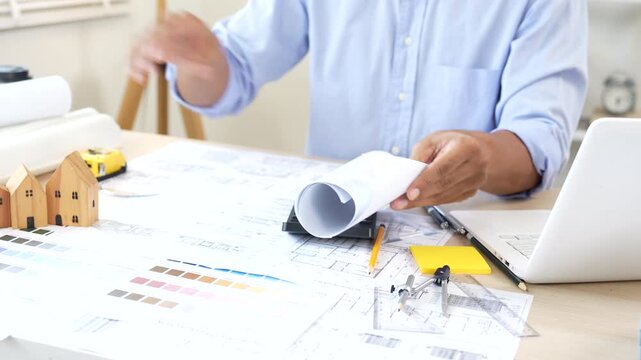 Architect reviewing construction plans at a modern office desk with blueprints, laptop, and engineering tools for a building project.