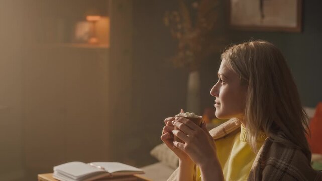 Caucasian young adult woman wrapped in blanket drinking hot cocoa with marshmallows at home. Footage is suitable for mental health, self-care, and seasonal wellness promotions