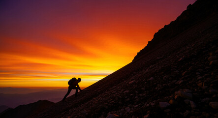 Fototapeta premium Silhouette of a hiker climbing a steep mountain during a vibrant sunset. Mountaineer ascending rocky terrain against a fiery orange sky. Success and determination concept for adventure marketing.