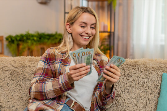 Young adult woman counts dollar bills on home sofa, smiling proudly and nodding after salary raise or jackpot win. Blonde girl manages family budget, enjoying moment of financial success and optimism.