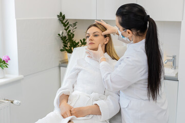 Middle-aged woman sits in chair while cosmetologist in gloves gently checks facial skin before treatment. Calm adult client in white robe enjoys skincare consultation inside modern beauty clinic room.