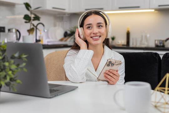 Young woman at home wears wireless headphones types message on smartphone takes break from tasks. Caucasian girl on sofa sits at table with laptop checks social app replies to chat relaxes happy mood.
