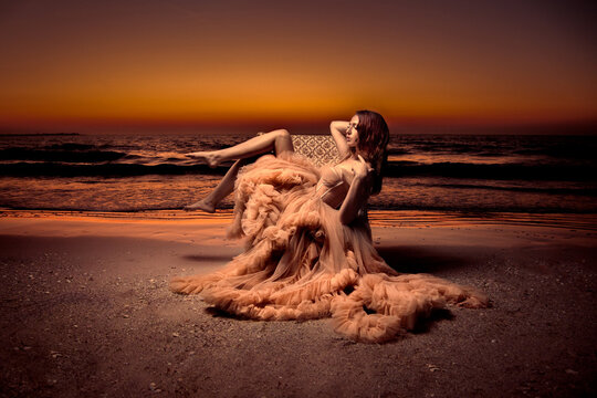 Side view of a Young woman in a full length evening gown sitting on a chair on a sandy beach at sunset, Florida, USA