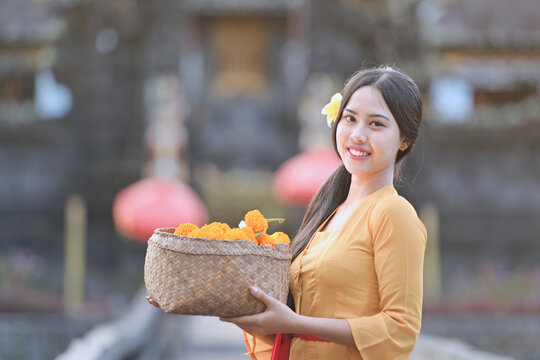 Portrait of a smiling Balinese woman in a traditional kebaya batik dress standing in front of a temple holding a basket of flowers, Indonesia