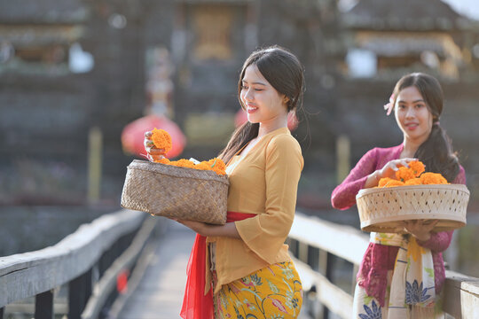 Portrait of two smiling Balinese women in a traditional kebaya batik dress standing in front of a temple holding baskets of flowers, Indonesia
