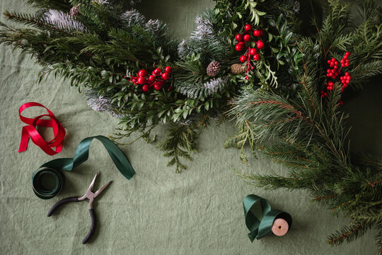 Overhead view of spools of ribbon, pliers and assorted foliage including berries, pinecones, fir branches on a table for making a Christmas wreath