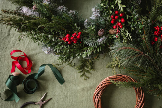 Overhead view of a Christmas wreath, ribbon, pliers and assorted foliage including berries, pinecones, fir branches on a table