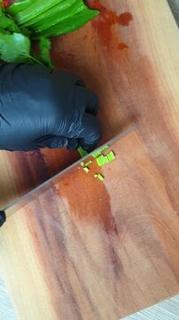 Chef wearing black gloves finely chopping fresh cilantro leaves on a wooden cutting board with a sharp knife in the kitchen