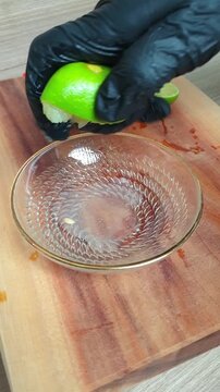 A hand in a black glove squeezing fresh lime juice into a clear glass bowl on a wooden cutting board