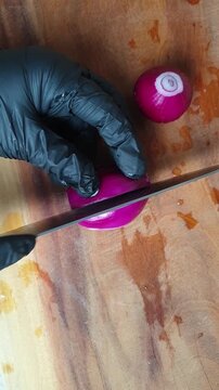 A chef wearing a black glove slicing a fresh red onion on a wooden cutting board with a sharp knife