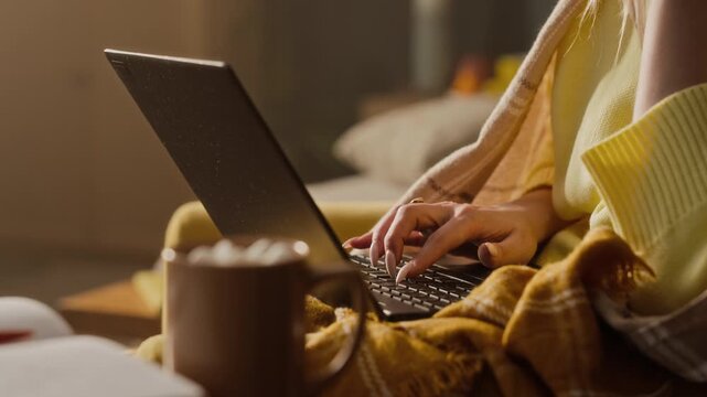 Young adult Caucasian woman wrapped in blanket typing on laptop and drinking hot beverage from mug. Footage is ideal for remote work, online learning, or seasonal wellness marketing campaigns