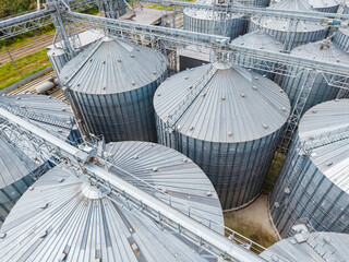 Large metal storage silos are arranged in rows at a grain facility during daylight hours in a rural area © DSMT