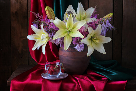 lilies and purple flowers sit in a clay vase on red silk fabric beside raspberries. Floral arrangement, classic elegance, and still life concept.