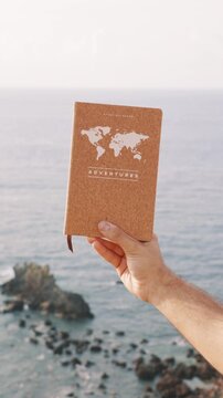vertical Male hand holding an adventure travel journal over the sea