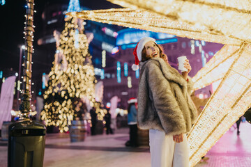 Fototapeta premium Woman wearing a Santa hat and fur coat smiles while holding a warm drink amid holiday lights. Cheerful scene with illuminated decorations and a Christmas tree at a nighttime market.